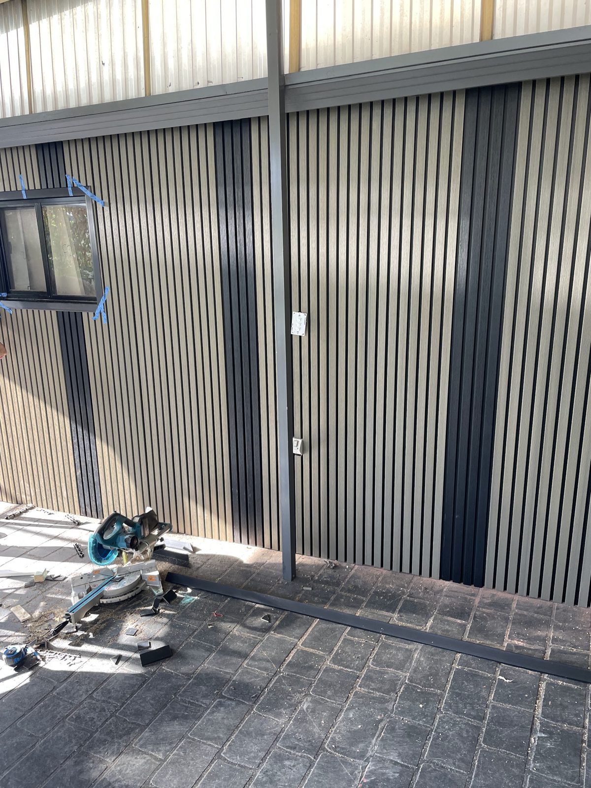 Vertical slat screening panels being fitted to an exterior wall, mitre saw on the paving
